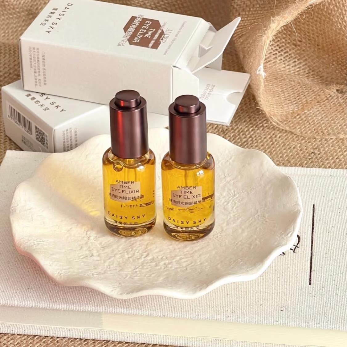 Two amber glass bottles with brown caps labeled Daisy Sky Amber Time Eye Elixir Oil on a shell dish with packaging